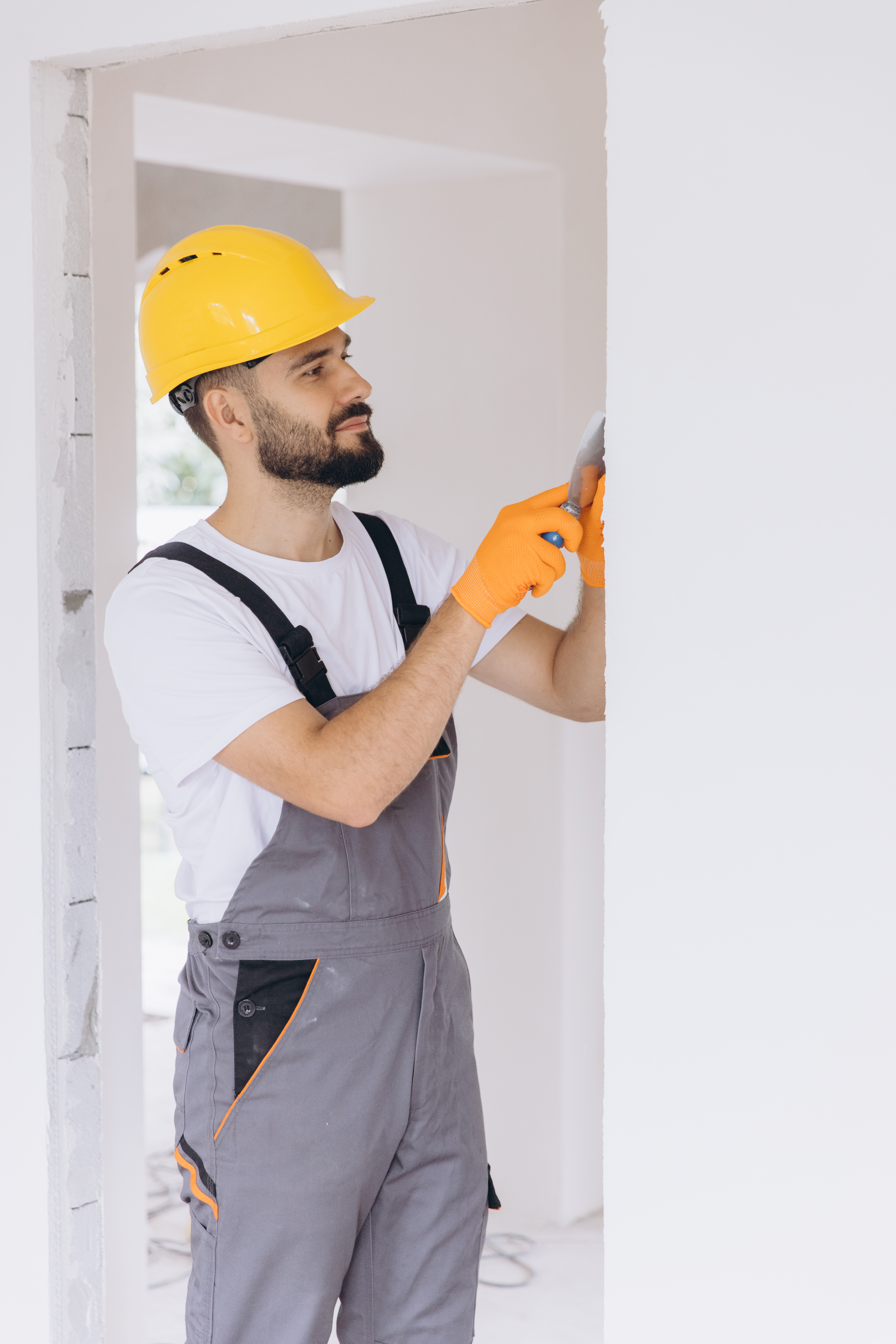 Construction worker in safety helmet and overalls applying spackle or plaster to a wall with a spatula tool, renovating a room