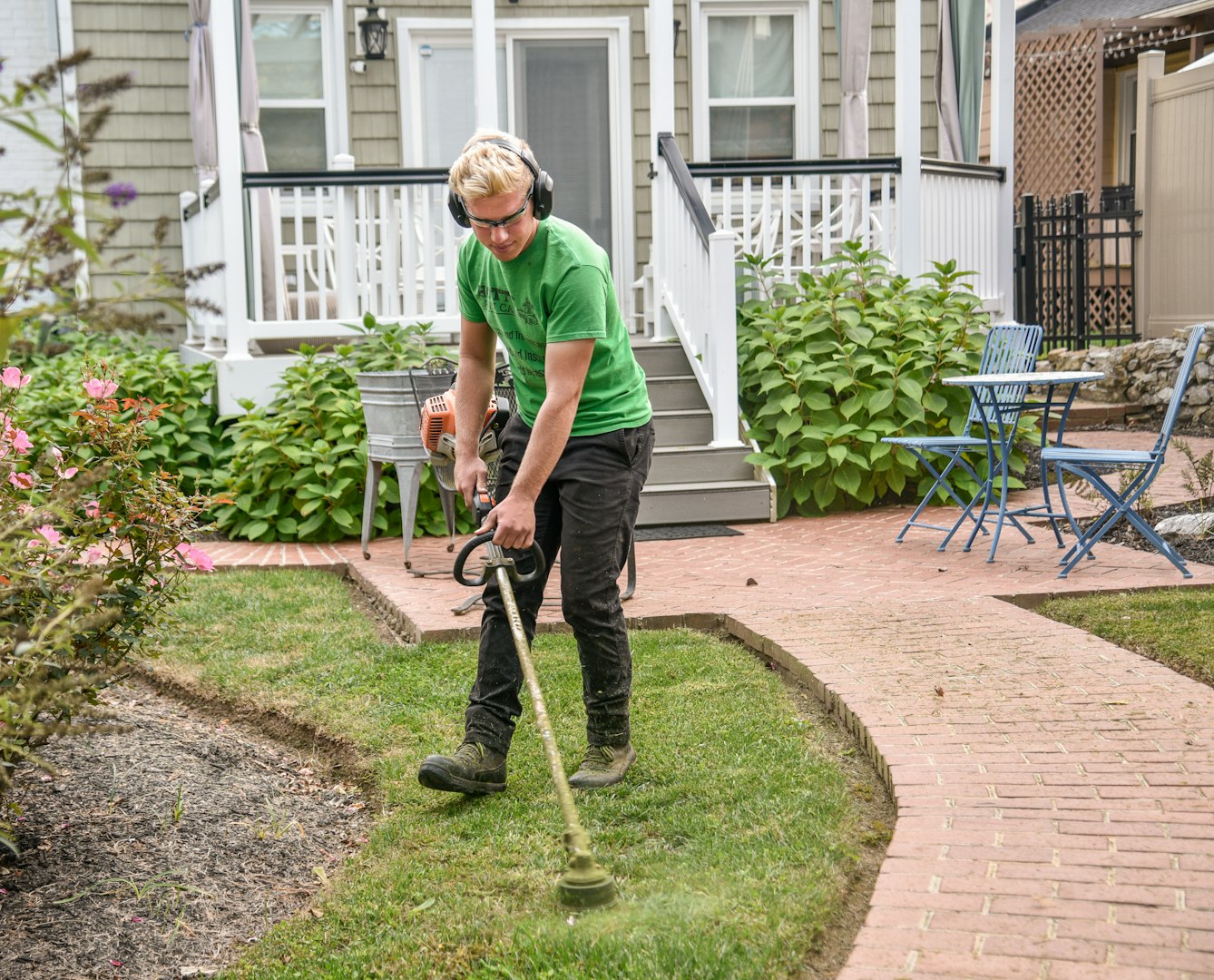 Man wearing headphones gathering green trimmings and cut branches from the garden path after pruning shrubs